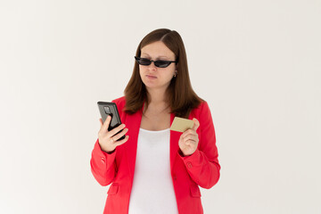 Cheerful young girl in black sunglasses showing plastic credit card while using mobile phone isolated over white background, wearing red jacket