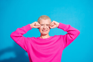 Naklejka premium A young woman in a bright pink sweater, squeezing her eyes in a playful gesture, stands against a vivid blue background
