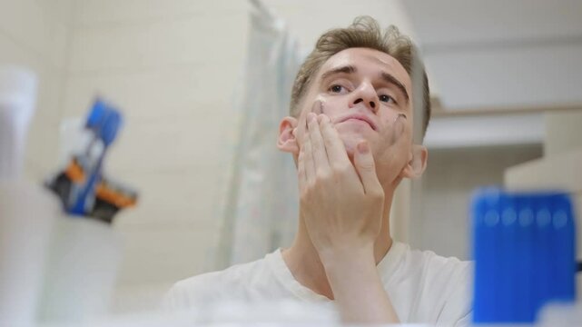 Young guy applies face wash or mask to face in front of mirror