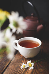 Cup of red tea, two chamomile flowers and kettle on wooden table. Blurred bouquet on foreground