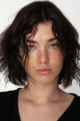Close-up of a young woman with wet-look wavy black hair and green eyes, wearing a black top, with a soft neutral background.