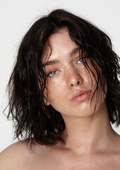 Minimalist portrait of a young woman with wavy black hair and subtle freckles, gazing directly at the camera, set against a bright white background