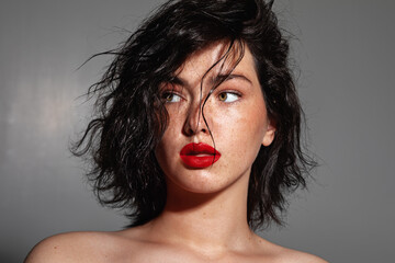  Portrait of a young woman with curly black hair, bold red lips, and natural freckles, looking to the side against a neutral gray background.