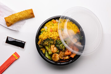 Chinese takeout featuring shrimp in garlic sauce with vegetables, served alongside fried rice, a spring roll, metal chopsticks, and a packet of soy sauce.