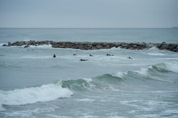 Surfers waiting for the perfect wave on a cloudy day. The breakwater in the background adds an interesting element to this dynamic scene, ideal for sports, travel, and lifestyle themes