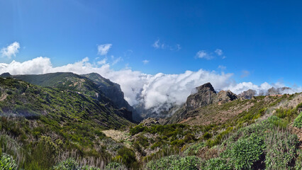 Breathtaking view of Vereda Pico do Arieiro in Madeira, Portugal, featuring vibrant green landscapes, dramatic mountain peaks, and fluffy clouds rolling through the valleys under a bright blue sky