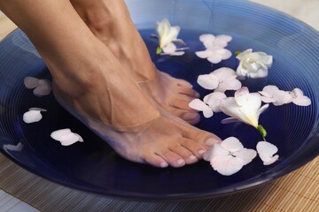 Woman soaking her feet in bowl with water and flowers on floor, closeup. Spa treatment