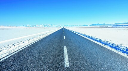 Fototapeta premium Empty asphalt road through snowy landscape leading to distant mountains under clear blue sky.
