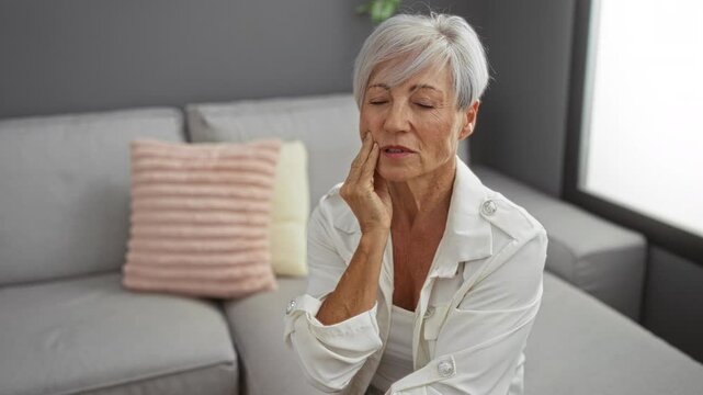 Elderly woman experiencing tooth pain while sitting in a modern living room with grey walls and a comfortable sofa adorned with a pink cushion in her home.