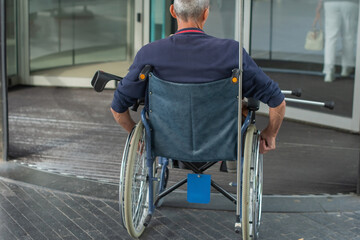 Rear view of a person in a wheelchair entering a building through automatic glass doors, urban...