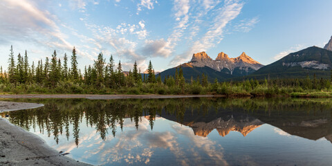 Three Sisters in Canmore seen at golden hour, sunset on blue sky day, afternoon with calm, peaceful reflection in water below famous, tourist, tourism mountains, area summer.	