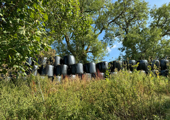 Stacks of black-wrapped hay bales are situated in a lush,grown area, surrounded by tall grasses and leafy trees. Sunlight filters through the leaves, casting  shadows near, Elslack, UK.  