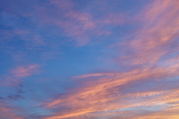 Fiery light orange hues with traces of pink and purple decorate streaky clouds drifting through a transitioning blue sky at dusk, and cover a crescent moon in a thin pink veil.