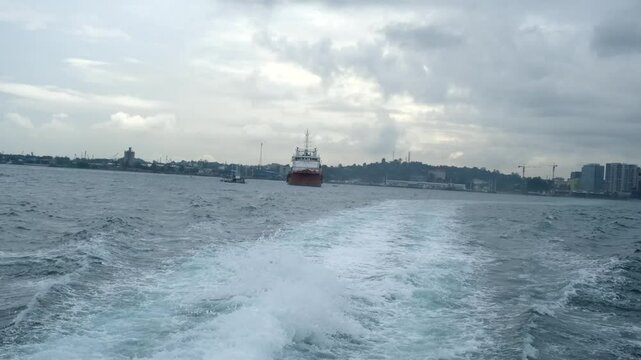 A red vessel sails near the Batam city skyline under a cloudy sky, leaving a foamy wake in the choppy sea.