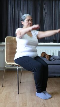 Senior woman with arms outstretched, doing arm stretching exercises on a chair, dachshund in the background. Concept of senior home workouts and healthy lifestyle