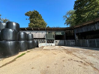 A farmyard scene shows a stack of large black hay bales beside a wooden barn. Metal gates stand...
