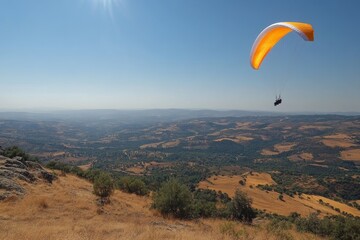 A paraglider soaring over a scenic landscape of hills and valleys under a clear blue sky.