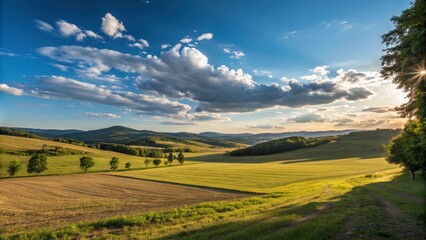 Obraz premium Rolling hills, bathed in golden sunlight, create a picturesque landscape, with vibrant green fields and a blue sky dotted with fluffy clouds.