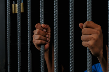 hands of a prisoner behind prison bars on black background	