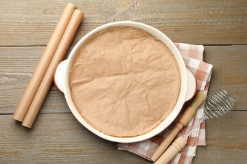 Ceramic casserole, baking parchment paper, brush and whisk on wooden table, flat lay