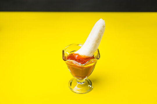 A banana piece dipped into a glass container of ketchup on a yellow and black background
