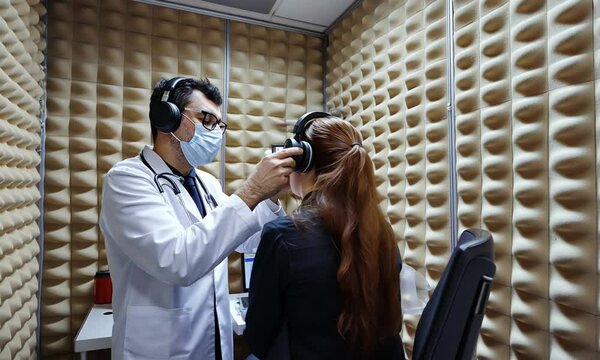 A medical professional adjusts headphones on a patient in a soundproof testing room.