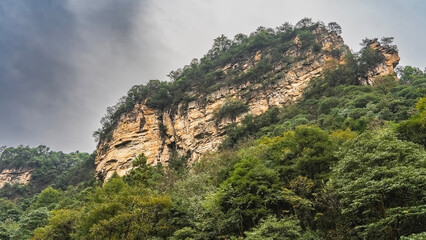 Naklejka premium A picturesque mountain against a cloudy sky. Steep rocky slopes. Lush green vegetation at the foot. China
