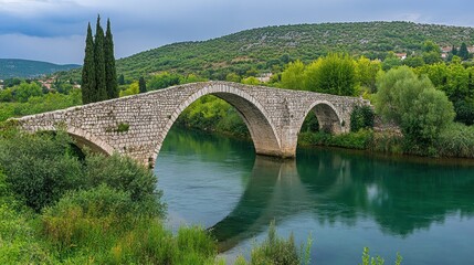 Fototapeta premium Ancient stone arch bridge over calm river, reflecting in water, surrounded by lush green vegetation and hills under a cloudy sky.