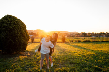 Teenage son giving mother piggyback ride at sunset in scenic grassy field