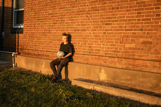 Boy with rugby ball sitting against brick wall in warm afternoon light