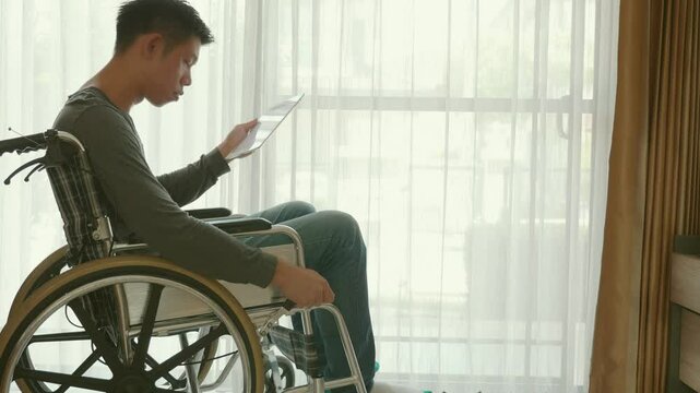 A man in a wheelchair is sitting in front of a window, looking at his cell phone. Concept of isolation and loneliness, as the man is alone in his wheelchair and not interacting with anyone else
