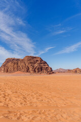 A stunning desert landscape featuring orange sand dunes, rugged rocky outcrops, and a vibrant blue sky adorned with delicate white clouds, showcasing the beauty and vastness of desert wilderness...