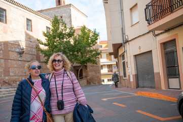 Two middle-aged women tourists smiling as they walk through a sunny street in a charming Spanish town, enjoying their travel experience near historic buildings