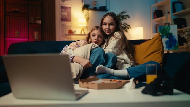 Teen girls watch a scary movie on a laptop, sitting on the sofa and hugging