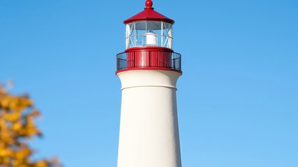 A tall white lighthouse with a red top against a clear blue sky, surrounded by autumn foliage, serving as a beacon for navigation.