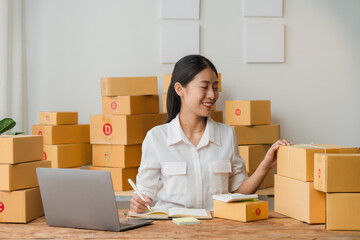 E-commerce Success: A young Asian woman, surrounded by a stack of shipping boxes, smiles confidently as she manages orders on her laptop.