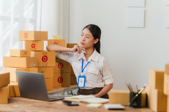 Entrepreneurial Woman Planning her Business: A young Asian businesswoman thoughtfully leans against a stack of shipping boxes in her home office, strategizing her next move.  Laptop, notebook. - Powered by Adobe