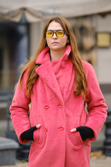 Fototapeta premium A young woman in a pink coat and yellow sunglasses stands confidently outdoors in a city setting
