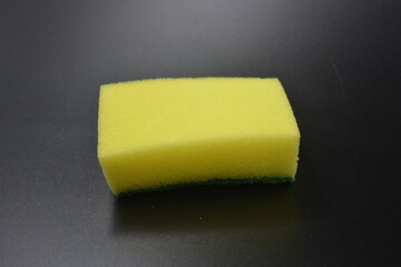 A small yellow sponge with a green rough dishwashing surface placed on a black, matte plastic background.