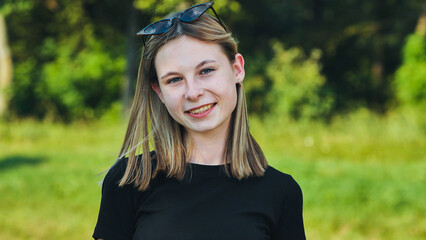 Teenage girl with blonde hair and sunglasses is smiling outdoors with a green background.