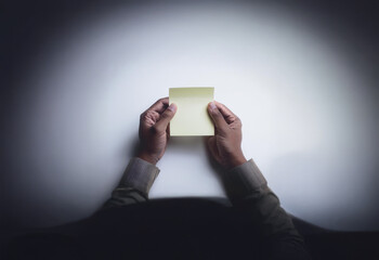 A pair of hands hold a blank yellow sticky note in a dimly lit room, highlighting the note as a symbol of potential.