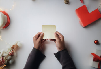 Obraz premium A pair of hands hold a blank yellow sticky note on a white table with festive Christmas decorations.
