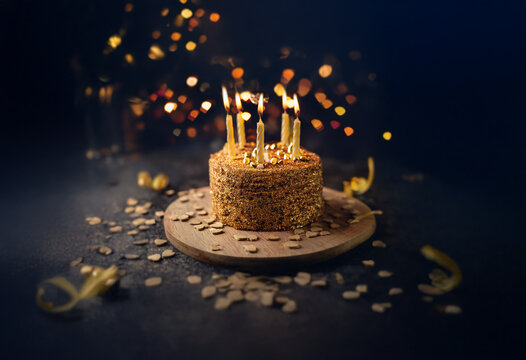 A birthday cake with lit candles sits on a wooden board against a dark background with bokeh lights.