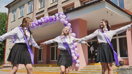 Three joyful graduates donning purple ribbons embrace their last day of school with open arms and smiles in front of the school