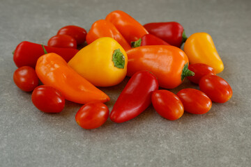 A vibrant assortment of mini sweet peppers in red, orange, and yellow hues alongside fresh red cherry tomatoes arranged on a gray stone background. 