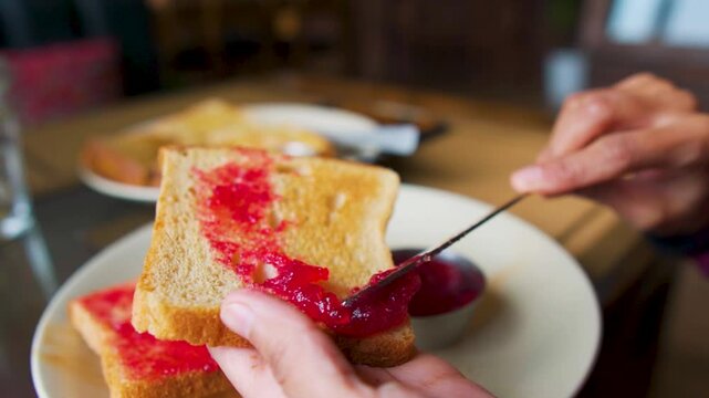 Close up shot of girl holding bread and applying jam on it. Hand holding knife spreading jam on toast. Morning breakfast.