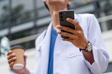 Male Doctor in Professional Uniform Holding Coffee and Smartphone Outdoors in Urban Setting
