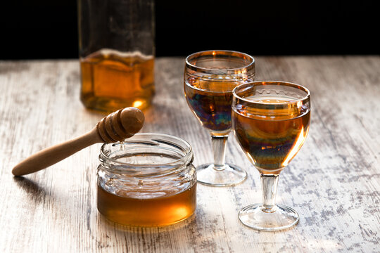 Still life close-up of two glasses of honey mead (medovukha) with honey dipper on wooden surface, dramatic side light