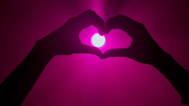 Black hand silhouette, person showing gesture and making heart sign against bright pink colored spotlight, isolated on studio background close up shot.