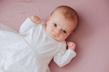 A baby is laying on a pink surface with a white dress on. The baby has a serious expression on its face